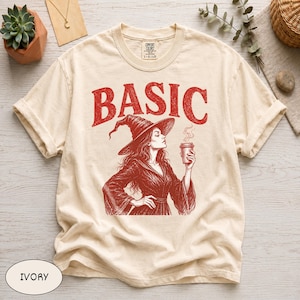 Può includere: Una t-shirt a maniche corte color avorio con una stampa grafica rossa raffigurante una strega con un cappello a punta che tiene in mano una tazza di caffè. La parola BASIC è stampata in grandi lettere rosse sopra l'illustrazione. Girocollo ed etichetta Comfort Colors.