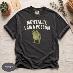 May include: A charcoal gray short-sleeve t-shirt featuring the text "MENTALLY, I AM A POSSUM" in white capital letters above a graphic illustration of a green toad with a tired expression. The shirt has a crew neckline and a vintage-washed finish.