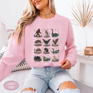May include: A light pink long-sleeve sweatshirt featuring a grid of twelve illustrated mythical creatures, including Bigfoot, a mothman, and a dragon, with each creature labeled in small text below its respective graphic illustration.