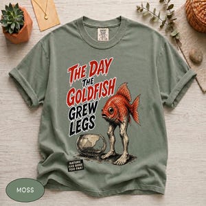 Könnte beinhalten: Ein salbeigrünes Kurzarm-T-Shirt mit einer Grafik eines Goldfisches mit menschlichen Beinen, der neben einem zerbrochenen Goldfischglas steht. Der Text lautet THE DAY THE GOLDFISH GREW LEGS und NATURE HAS GONE TOO FAR. Rundhalsausschnitt.