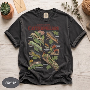 May include: A charcoal gray short-sleeve t-shirt featuring a graphic print titled Field Guide to Caterpillars. The design displays various illustrated caterpillars with their names, including Monarch, Tomato Hornworm, and Hickory Horned Devil.