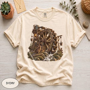May include: An ivory short-sleeve t-shirt featuring a graphic print of a brown beaver standing near a large wooden water wheel. The scene includes a rustic structure with hanging buckets, flowing water, and mossy rocks. The shirt has a crew neckline.