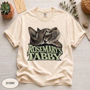 May include: An ivory short-sleeve t-shirt featuring a graphic of a brown tabby cat looking upward. Below the cat, the text reads ROSEMARY'S TABBY in large, bold, light green letters. The shirt has a crew neckline and a Comfort Colors brand label.