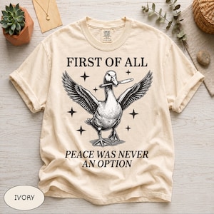Peut inclure: T-shirt ivoire avec le texte "FIRST OF ALL" au-dessus d'une illustration détaillée d'un canard aux ailes déployées, tenant un couteau. En dessous du canard, le texte se lit "PEACE WAS NEVER AN OPTION".