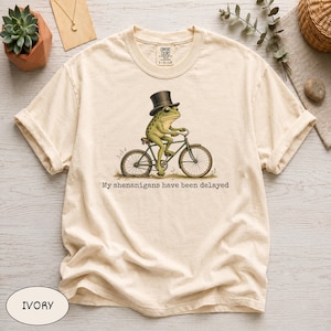 Könnte beinhalten: Ein elfenbeinfarbenes Kurzarm-T-Shirt mit einer Grafik eines grünen Frosches, der einen schwarzen Zylinder trägt und Fahrrad fährt. Unter der Illustration steht der Text, My shenanigans have been delayed. Das Shirt hat einen Rundhalsausschnitt.