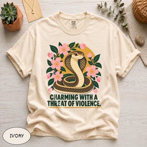 May include: An ivory t-shirt featuring a graphic of a coiled cobra surrounded by pink flowers and green leaves against a yellow circle. Below the snake, the text reads: CHARMING WITH A THREAT OF VIOLENCE. The shirt has a crew neckline and short sleeves.