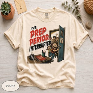 以下が含まれることがあります： THE PREP PERIOD INTERRUPTEDというテキストがプリントされたアイボリーのTシャツ。イラストには、子供たちが入ってくる教室のドア、FIVE MORE MINUTESと書かれたマグカップ、LESSON PLANSと書かれた本が置かれた机が描かれています。