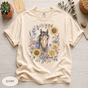 Könnte beinhalten: Ein elfenbeinfarbenes Kurzarm-T-Shirt mit einer zentralen Grafik eines braunen Pferdekopfes, umgeben von einem farbenfrohen Kranz aus Sonnenblumen, Rosen und Lavendelzweigen. Das Shirt hat einen Rundhalsausschnitt und ein Comfort Colors Etikett.