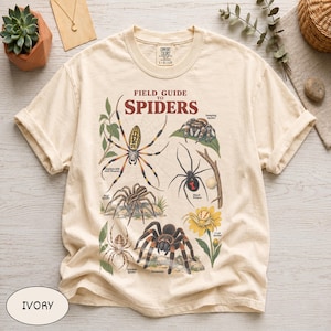 Puede incluir: Una camiseta de manga corta color marfil con un estampado gráfico titulado Field Guide to Spiders. El diseño muestra ilustraciones de varias arañas, incluyendo una araña tejedora de seda dorada, una saltarina, una viuda negra y una tarántula.