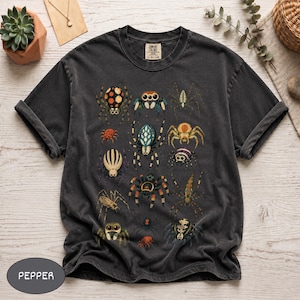 May include: A charcoal grey short-sleeve t-shirt featuring a graphic print of various colourful spiders in different shapes and sizes. The shirt has a crew neckline and a Comfort Colors brand label on the inside collar. The word PEPPER is printed.