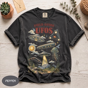 Könnte beinhalten: Ein anthrazitfarbenes T-Shirt mit einem Grafikdruck mit der Aufschrift FIELD GUIDE UFOS. Das Design zeigt verschiedene fliegende Untertassen, eine leuchtende Sonne und ein quallenartiges Raumschiff über einer Waldlandschaft mit Bergen.