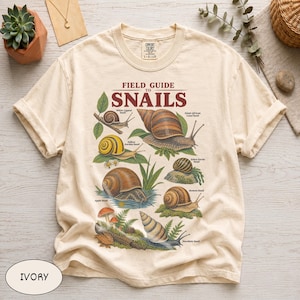 May include: An ivory short-sleeve t-shirt featuring a graphic print titled Field Guide to Snails. The design displays various snail species including the White-Lipped, Giant African Land, Yellow Garden, Zebra Nerite, Apple, and Roman snails with foliage.