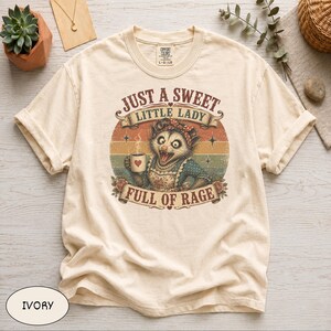 以下が含まれることがあります： アイボリー色のTシャツ。マグカップを持ったオポッサムのヴィンテージ風グラフィック。テキストは「JUST A SWEET LITTLE LADY FULL OF RAGE」と書かれています。Comfort Colorsブランド、サイズL-G/LGのシャツです。