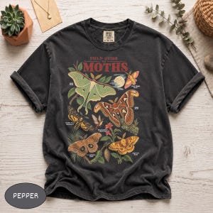 Könnte beinhalten: Ein anthrazitfarbenes T-Shirt mit einem Grafikdruck mit dem Titel Field Guide to Moths. Das Design zeigt verschiedene Mottenarten in Grün-, Braun- und Gelbtönen, begleitet von kleinen Textbeschriftungen für jedes Insekt auf einem dunklen Hintergrund.
