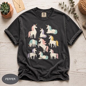 Può includere: Una t-shirt a maniche corte grigio antracite con una stampa grafica di otto unicorni in varie pose. Gli unicorni hanno criniere e code color pastello nelle tonalità del rosa, blu e giallo. La maglietta ha un girocollo e un'etichetta sul collo.