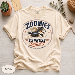 May include: An ivory t-shirt featuring a graphic of a dog wearing a harness and carrying a cardboard box. The text reads "ZOOMIES EXPRESS Delivery" and "FAST FOR ABSOLUTELY NO REASON." The design is enclosed in a circular frame with red stars.