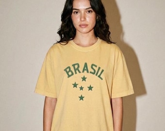 Könnte beinhalten: Ein gelbes T-Shirt mit dem Wort "BRASIL" in grünen Buchstaben und vier grünen Sternen. Das T-Shirt hat kurze Ärmel und einen Rundhalsausschnitt. Das Model trägt das T-Shirt.
