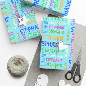 Può includere: Due regali rettangolari avvolti in carta azzurra con il nome Stephanie stampato ripetutamente in vari caratteri, dimensioni e colori, tra cui arancione, verde, blu e rosa. Ogni regalo è legato con spago marrone e un'etichetta bianca.