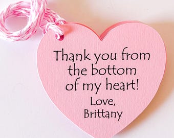 以下が含まれることがあります： 「Thank you from the bottom of my heart! Love, Brittany」のテキストが入ったピンク色のハート型のギフトタグ。ピンクと白の紐が付いています。タグは紙製です。