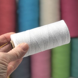 May include: A white cotton twine spool, held in a hand, against a blurred background of colorful spools of thread.