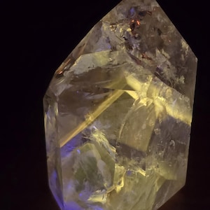 May include: A clear, multifaceted quartz crystal point with a pointed top and a hexagonal base. The crystal features internal fractures and inclusions that catch the light, displaying translucent white and yellow tones with a subtle blue glow.