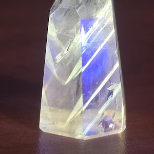 May include: A clear, faceted quartz crystal point with a pointed top and a flat base. The crystal features internal fractures and inclusions that catch the light, displaying a distinct, vibrant blue glow within its transparent structure.