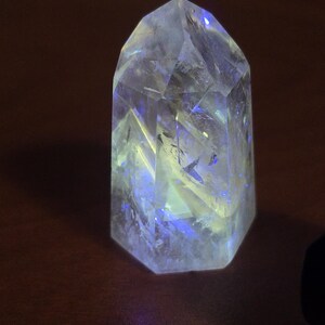 May include: A clear, faceted crystal point with a hexagonal base. The translucent stone features internal fractures and inclusions that catch the light, displaying vibrant blue and yellow highlights throughout its structure.