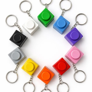 May include: A collection of colorful square keychains in various colors, including green, blue, purple, pink, red, orange, yellow, gray, white, and black. Each keychain features a circular button on the top and a silver keyring.
