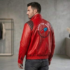 Op de afbeelding: Rode leren jas met bruine schouderstukken. De achterkant toont een afbeelding van een rood-blauwe pil met de tekst "GOOD FOR HEALTH BAD FOR EDUCATION". De jas heeft een bomberstijl met een geribbelde zoom en manchetten.