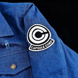 May include: Blue denim jacket with a Capsule Corp patch on the upper sleeve. The patch is white with a black circular design and the words "CAPSULE CORP" in black. The jacket has a gold button and a distressed area near the button.