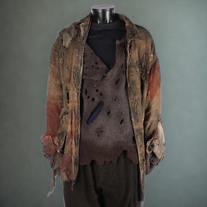 May include: A distressed costume ensemble on a mannequin. The outfit includes a tattered brown jacket with red accents, a dark gray vest with holes, and dark pants. The clothing appears worn and aged, suggesting a historical or theatrical theme.
