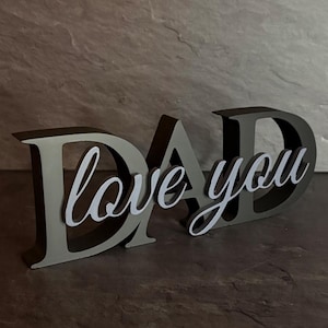 Könnte beinhalten: Ein dunkelgraues, dreidimensionales, freistehendes Schild, das das Wort DAD in Großbuchstaben buchstabiert. Der Schriftzug love you ist in weißer Schreibschrift auf der Vorderseite der Buchstaben angebracht und bildet ein mehrschichtiges Objekt.