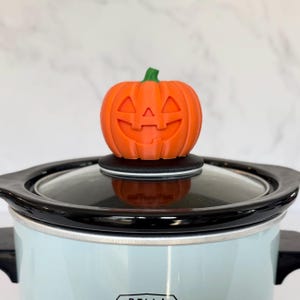 May include: An orange pumpkin-shaped lid knob with a green stem sits atop a slow cooker. The pumpkin has carved-out eyes, nose, and a smiling mouth. The slow cooker is light blue with a black rim and handles.