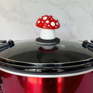 May include: A red slow cooker with a glass lid and a decorative mushroom-shaped knob. The mushroom has a red cap with white spots and a white stem. The slow cooker is red.
