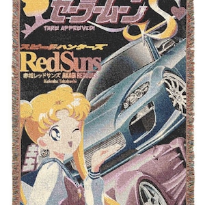 Könnte beinhalten: Gewebte Überwurfdecke mit einem Cartoon-Bild einer Sailor Moon-Figur und einem blauen Auto, mit dem Text "Red Suns" und japanischem Text. Die Decke hat einen schwarzen Hintergrund, Fransen und ein farbenfrohes Design.
