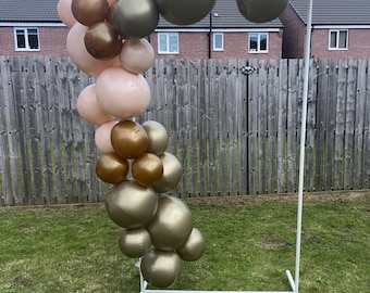May include: A balloon arch with a variety of colors, including gold, bronze, and blush pink. The balloons are arranged in a curved shape and attached to a white metal frame. The arch is set up outdoors on a grassy lawn.