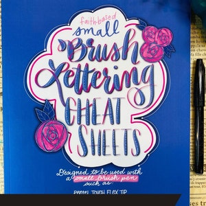 May include: A blue book with the title "Faith-based small brush lettering cheat sheets" in pink and blue lettering. The book is designed to be used with a small brush pen, such as a Pentel Touch Flex Tip. The book is shown with a pen and a book/pen bundle label.