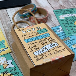 May include: A brown paper gift bag with a yellow and blue card attached. The card has the text "The Lord is my light and my salvation - whom shall I fear? The Lord is the stronghold of my life - of whom shall I be afraid?"  The bag is decorated with a pink and white gingham ribbon.