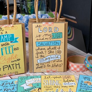 May include: Four small brown paper bags with colorful printed cards inside. The cards feature bible verses with handwritten text in a variety of colors. The text on the cards includes "Your word is a lamp unto my feet and a light unto my path. Psalm 119:105", "The Lord is my light and my salvation - whom shall I fear? The Lord is the stronghold of my life - of whom shall I be afraid? Psalm 27:1", "Jesus said "Let your light shine before others, that they may see your good deeds", and "You are the light of the world. A city set on a hill cannot be hidden, nor do they light a lamp and put it under a bushel, but on a stand, and it gives light to all in the house. Matthew 5:14-16".