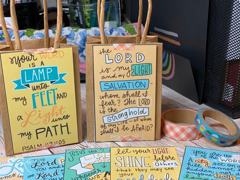 May include: Four small brown paper bags with colorful printed cards inside. The cards feature bible verses with handwritten text in a variety of colors. The text on the cards includes "Your word is a lamp unto my feet and a light unto my path. Psalm 119:105", "The Lord is my light and my salvation - whom shall I fear? The Lord is the stronghold of my life - of whom shall I be afraid? Psalm 27:1", "Jesus said "Let your light shine before others, that they may see your good deeds", and "You are the light of the world. A city set on a hill cannot be hidden, nor do they light a lamp and put it under a bushel, but on a stand, and it gives light to all in the house. Matthew 5:14-16".