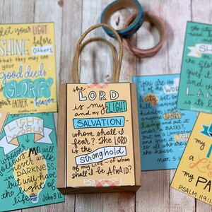 May include: A set of colorful cards with inspirational quotes about light and faith. The cards are arranged around a small brown paper bag with a yellow and orange design. The bag has the text "The Lord is my light and my salvation" on it.