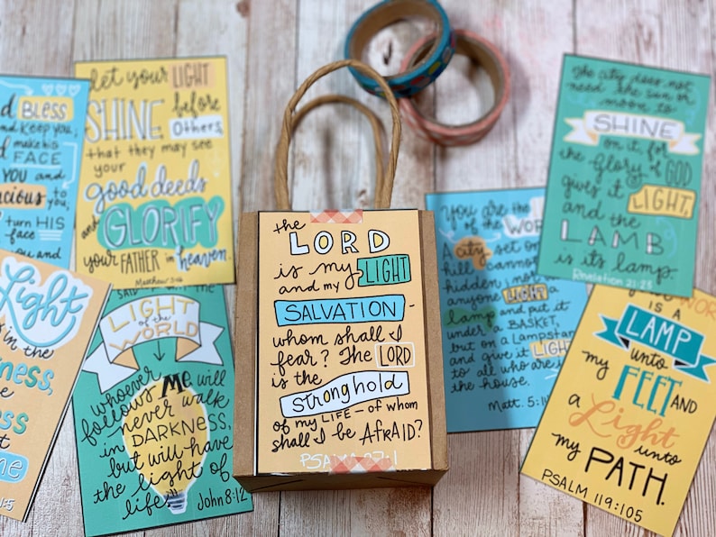May include: A set of colorful cards with inspirational quotes about light and faith. The cards are arranged around a small brown paper bag with a yellow and orange design. The bag has the text "The Lord is my light and my salvation" on it.