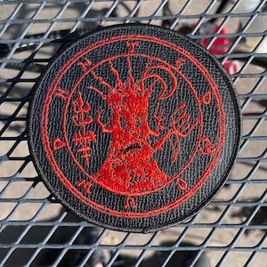 May include: Round black patch with a red embroidered design. The design features a central figure with a face and a surrounding circular border with text. The patch is likely for clothing or accessories.