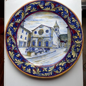 Può includere: Un piatto decorativo in ceramica con una pittura dettagliata di una chiesa e di una piazza. Il piatto ha un bordo floreale blu, rosso e giallo. Il piatto è rotondo e ha un bordo dorato. Il testo "Nedo da Gualdo" è visibile.