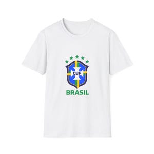 Puede incluir: Camiseta blanca con el logo de la Confederación Brasileña de Fútbol (CBF). El logo tiene forma de escudo con detalles azules, amarillos y verdes, las letras "CBF" y la palabra "BRASIL" en verde.