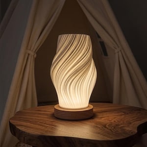 May include: A white, swirling ceramic lamp with a wooden base sits on a dark wooden table. The lamp's design features a textured, wave-like pattern. The background includes a light-colored tent.