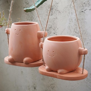 May include: Two terracotta-colored hanging planters with smiling faces and arms, suspended by rope. The planters have a whimsical design, with a round body and a small, attached base. The planters are designed to hold plants.
