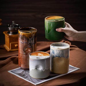 May include: Four ceramic coffee mugs in shades of brown, green, blue, and gray. The mugs have a unique design with a small opening at the top. One mug is being held by a hand. A wooden coffee grinder is in the background.