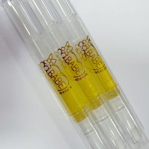 May include: Three clear nail oil pens with transparent caps. The pens contain a yellow liquid and gold floral accents with the word "BABE". The pens are angled diagonally against a white background.