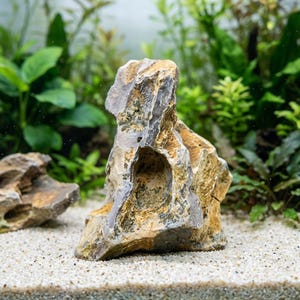 May include: A textured, multi-toned rock with a hole in the center, suitable for aquarium decoration. The rock features shades of gray, tan, and yellow, and is placed on a bed of white sand. Green plants are visible in the background.